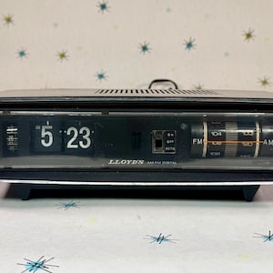 Puede incluir: Un radio reloj digital AM/FM Lloyd's negro y plateado vintage. El reloj muestra las 5:23 con una pantalla tipo flip. El dial de la radio es visible y la unidad tiene un asa en la parte superior. El radio reloj se encuentra sobre una base negra.