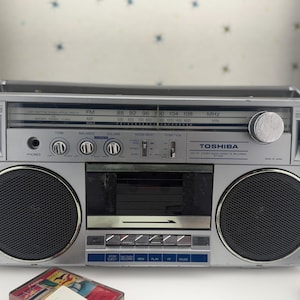 Op de afbeelding: Een zilverkleurige Toshiba stereo cassette recorder met radio-tuner voor FM- en AM-frequenties, een centraal cassettedeck, twee ronde luidsprekers, een hoofdtelefoonaansluiting en diverse bedieningsknoppen voor toon, balans en volume.
