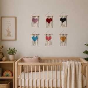 May include: Six small macrame wall hangings featuring heart-shaped designs in purple, red, black, blue, pink, and yellow. Each piece is crafted from off-white cord, suspended from a wooden dowel with a hanging string, and finished with fringe.