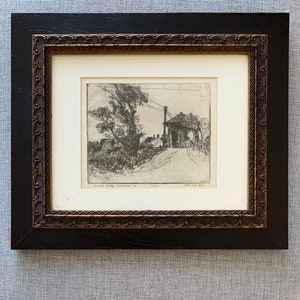 May include: A black and white etching of a bridge with a road leading to it. The etching is framed in a black frame with a gold trim. The title of the etching is "Covered Bridge, Millbrook, N.Y." and the artist's name is "Schuler" and the date is "State, Cope, 1924".