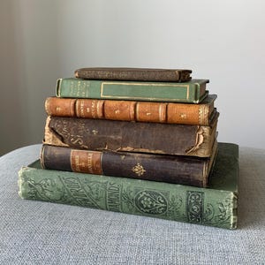 May include: A stack of antique books with aged covers in various colors, including green, brown, and tan. The books have visible wear and tear, suggesting they are vintage. The titles and authors are partially visible.