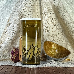 May include: A clear glass bottle with a gold lid and a gold design of branches on the side. The bottle is sitting on a wooden surface with a gold bowl and a purple rose.