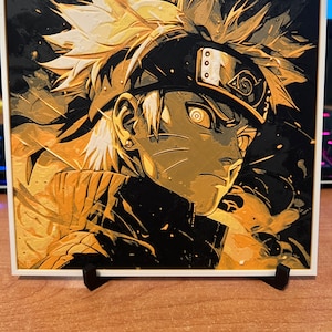 Può includere: Una stampa incorniciata di un'illustrazione stilizzata di un personaggio dell'anime Naruto. Il personaggio ha i capelli biondi a punta, una fascia per la testa con il simbolo "Konoha" su di essa e un'espressione seria. La stampa è in tonalità oro e nero.
