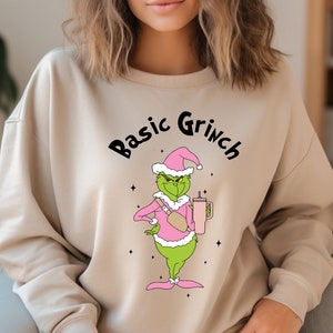 May include: A beige sweatshirt with the text "Basic Grinch" printed on the front. The Grinch is wearing a pink Santa hat and holding a pink cup.