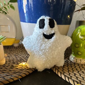 May include: White plush ghost with black eyes and a smiling mouth. The ghost is sitting on a woven mat with string lights.