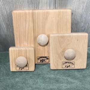 Peut inclure: Trois blocs de bois de différentes tailles, chacun avec un bouton arrondi. Les blocs sont brun clair avec un grain de bois visible. Les blocs les plus petits et les plus grands sont carrés, tandis que celui du milieu est rectangulaire. Chaque bloc a une petite étiquette rectangulaire avec du texte.