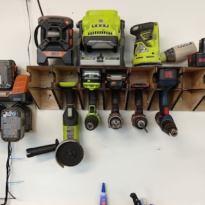 Może przedstawiać: Drewniany organizer ścienny z różnymi narzędziami elektrycznymi, w tym wiertarkami, szlifierką, szlifierką kątową i lampą roboczą. Organizer ma gniazda na każde narzędzie i stację ładowania baterii. Narzędzia są w różnych odcieniach zieleni, czerni i pomarańczy.