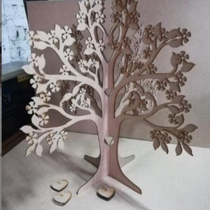 May include: A wooden tree cutout with a decorative design of flowers and leaves. The tree is freestanding and has a base with three legs. There are three small wooden hearts on the ground in front of the tree.