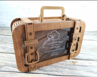 May include: A wooden suitcase-shaped money box with a clear plastic window. The window shows a logo with a plane and the text "ПУТЕШЕСТВИЕ  вещь, ПОКУПАЯ КОТОРУЮ БОГАЧЕ"