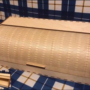May include: A wooden bread box with a hinged lid and a small slot for removing bread. The box is made of light brown wood and has a patterned design.