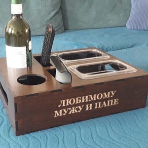 Puede incluir: Una bandeja de sofá de madera marrón oscuro con asas recortadas, que contiene una botella de vino, un mando a distancia y dos recipientes transparentes. La bandeja tiene el texto "Любимому мужу и папе" en la parte delantera.