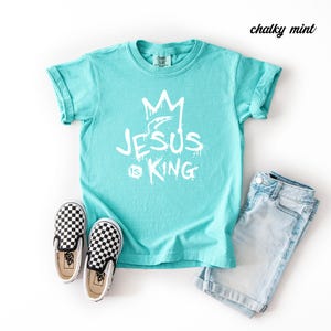 May include: A turquoise t-shirt with the text "JESUS IS KING" in white, paired with checkered slip-on sneakers and light blue denim shorts. The shirt has a crown graphic above the text.