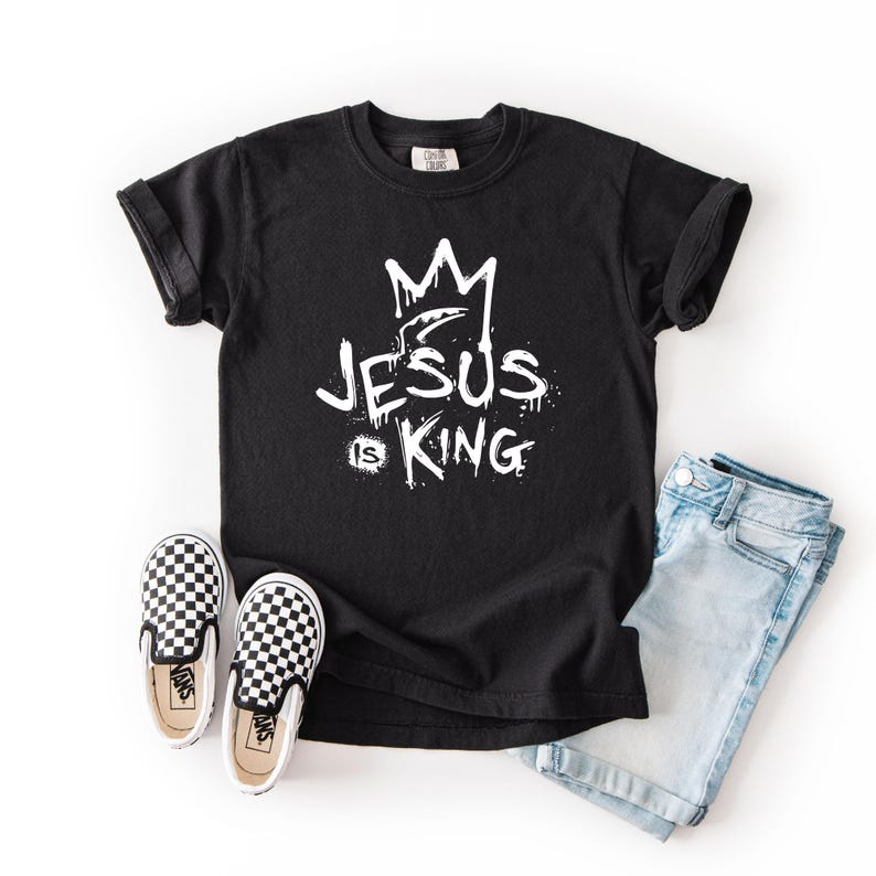 May include: Black t-shirt with the text "JESUS is KING" in white, along with a crown graphic. The shirt is paired with light blue denim jeans and black and white checkered slip-on shoes.