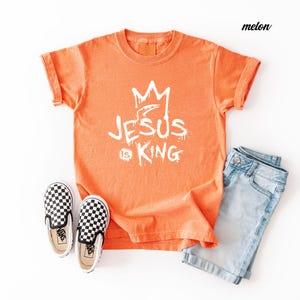 May include: An orange t-shirt with the text "JESUS is KING" in white, paired with light blue denim shorts and black and white checkered slip-on shoes. The shirt has rolled-up sleeves.