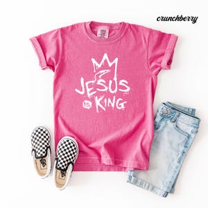 May include: A hot pink t-shirt with the text "JESUS IS KING" in white, along with a crown graphic. The shirt is paired with light blue denim shorts and black and white checkered slip-on shoes.