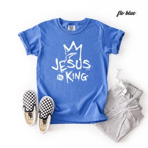 May include: A blue t-shirt with the text "JESUS is KING" in white, paired with gray sweatpants and black and white checkered slip-on shoes. The shirt has a crown graphic above the text.