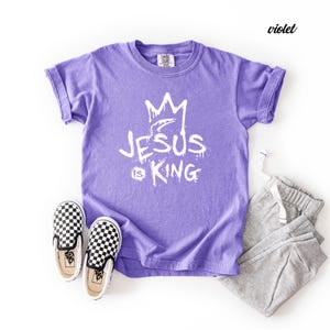 May include: A violet t-shirt with the text "JESUS IS KING" in white, paired with gray sweatpants and black and white checkered slip-on shoes. The t-shirt has rolled-up sleeves.