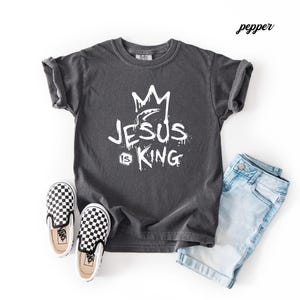 May include: A charcoal gray t-shirt with the text "JESUS IS KING" in white, featuring a crown graphic. The image also includes a pair of black and white checkered slip-on shoes and light blue denim shorts.