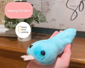 May include: A handmade, plush blue tadpole toy with black button eyes and a pink mouth. The toy is held in a hand. Text on the image reads "Sewing Pattern by apple seed sewing."