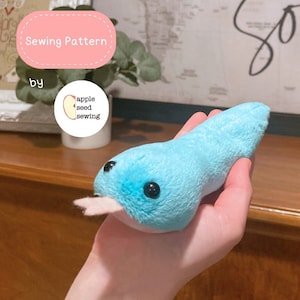May include: A handmade, plush blue tadpole toy with black button eyes and a pink mouth. The toy is held in a hand. Text on the image reads "Sewing Pattern by apple seed sewing."