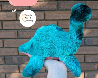 Könnte beinhalten: Ein blaugrünes Plüsch-Dinosaurierspielzeug mit langem Hals. Das Spielzeug wird in einer Hand gehalten. Der Text "Sewing Pattern by apple seed sewing" ist auf dem Bild zu sehen.