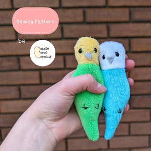 May include: Sewing pattern for two plush parakeets, one green and one blue, with yellow and white accents. The pattern is by apple seed sewing.