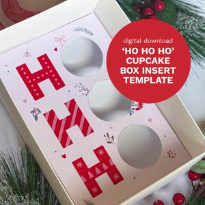 以下が含まれることがあります： 赤い「HO HO HO」の文字がプリントされたピンク色のインサートが入った白いカップケーキボックス。インサートにはカップケーキ用の円形の切り抜きがあり、装飾的な花柄とハートのアクセントが付いています。赤い円はデジタルダウンロードを示しています。