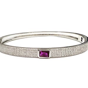 May include: A silver-toned hinged bangle bracelet featuring a pavé-set surface of small clear stones and a single rectangular, deep pink gemstone set in a silver-toned bezel at the center of the band, with a secure clasp mechanism on the side.