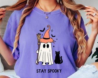 May include: A lilac-colored t-shirt featuring a Halloween-themed graphic. The design includes a ghost wearing a witch's hat and holding a broom, with a black cat and the text "STAY SPOOKY".