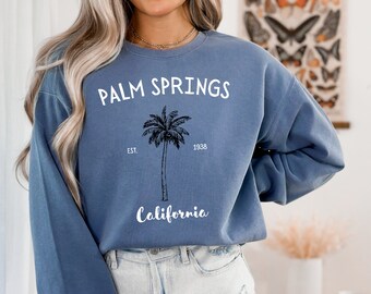 Puede incluir: Una sudadera azul con las palabras "PALM SPRINGS" en blanco, sobre una ilustración de palmera negra. Debajo, las palabras "California" y los números "1938" y "EST."
