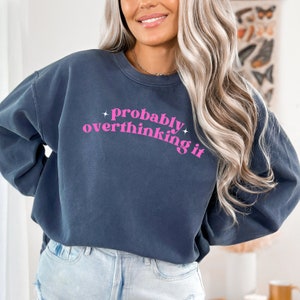 Puede incluir: Sudadera azul marino con la frase "probably overthinking it" en rosa. El texto está arqueado y tiene detalles de estrellas. La sudadera es de corte holgado.