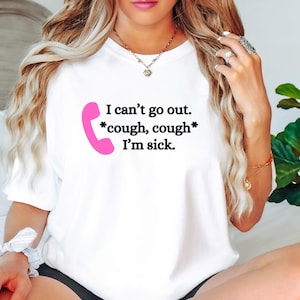 May include: White t-shirt with the text "I can't go out. *cough, cough* I'm sick." in black font. A pink telephone receiver graphic is on the left side of the text. The shirt is a casual style.