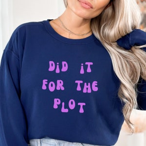 May include: A navy blue sweatshirt featuring the text "DID IT FOR THE PLOT" in purple, rounded lettering. The garment has a crew neck and is a solid colour.