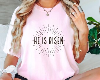 May include: A light pink t-shirt with the text "HE IS RISEN" in black, surrounded by a sunburst design. The shirt is a soft, casual style.