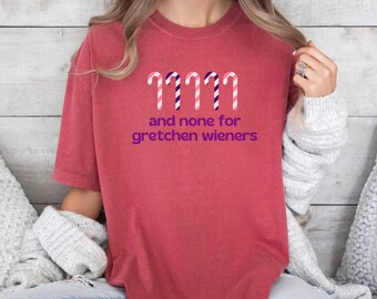 以下が含まれることがあります： 紫色の「and none for gretchen wieners」の文字が入った赤みがかったオレンジ色のTシャツ。テキストの上には、ピンクと紫のキャンディケーンが4つあります。カジュアルなスタイルのシャツです。