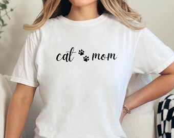 May include: White t-shirt with the words "cat mom" in black script, with paw prints between the words. The shirt is a casual style, suitable for everyday wear.