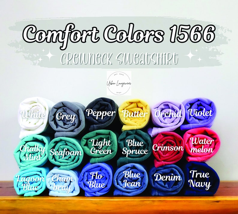 May include: A display of rolled-up Comfort Colors 1566 crewneck sweatshirts in various colors, including white, grey, pepper, butter, orchid, violet, chalky mint, seafoam, light green, blue spruce, crimson, watermelon, lagoon blue, chambray, flo blue, blue jean, denim, and true navy.