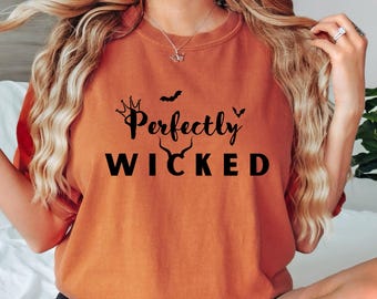 May include: Orange-colored t-shirt with the text "Perfectly Wicked" in black. The design includes a crown, bats, and horns, creating a Halloween or spooky theme. The shirt is a crew neck style.