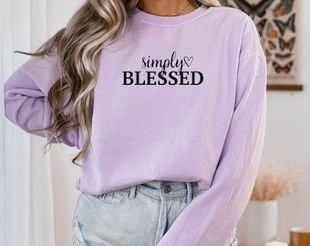 May include: A light purple sweatshirt with the words "simply BLESSED" in black script and block letters. The word "simply" is above the word "BLESSED" with a heart symbol. The sweatshirt is long-sleeved and has a crew neck.