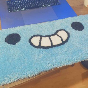Peut inclure: Un tapis rectangulaire bleu clair avec un motif de visage de dessin animé. Le tapis présente deux cercles noirs pour les yeux et une bouche souriante blanche et noire. Le tapis est fait d'une matière douce et moelleuse.