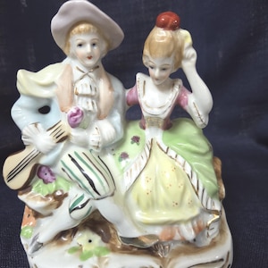 May include: A porcelain figurine depicting two figures seated together. The male figure wears a light blue coat, striped breeches, and a hat, holding a stringed instrument. The female figure wears a pink bodice and a green and yellow skirt.