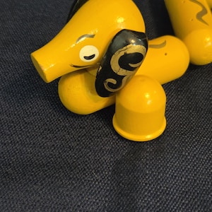 May include: A yellow wooden dachshund toy with black and gold accents on its ears. The toy has a simple design with a rounded body and a small, yellow tail. The dachshund is lying down on a dark blue surface.