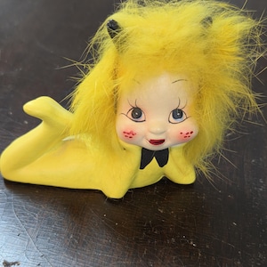 May include: A vintage ceramic figurine of a reclining figure with a yellow body and fluffy yellow hair. The figure has a painted face with rosy cheeks, blue eyes, and a black bow tie. The figurine is lying on a dark wooden surface.