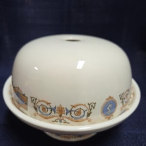 May include: A white ceramic cheese dome with a matching base. The dome has a small hole at the top. The base and dome are decorated with a repeating pattern of blue, orange, and green scrollwork and medallions. The cheese dome is on a dark blue surface.
