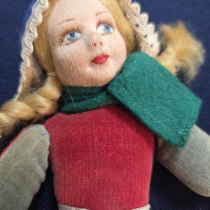 May include: A vintage doll with a fabric face, blonde plaited hair, and blue eyes. The doll is dressed in a red top, green scarf, and a white bonnet. The doll's arms and legs are grey and beige.