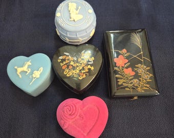 May include: Collection of decorative boxes in various shapes and colors. Includes a light blue round box with a cameo, a heart-shaped box with a unicorn, a black heart-shaped box with floral design, a black rectangular box with floral design, and a pink heart-shaped box.