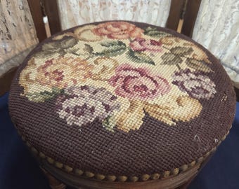 May include: A round footstool featuring a dark brown needlepoint top with a floral pattern of pink, yellow, and purple roses. The base is constructed from wood with a circular frame, decorative nailhead trim along the edge, and short legs.