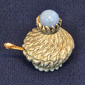 May include: A small, round, cream-colored container with a textured, swirling surface pattern and a smooth, light blue spherical knob on top. A thin, gold-toned metal spoon with a small, oval, light-colored inlay extends from the side.