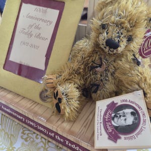 May include: A vintage-style teddy bear with a tag commemorating the 100th Anniversary (1902-2002) of the Teddy Bear. The bear is light brown with a black nose and paw pads. A framed print with the same text is also visible.