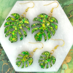 Könnte beinhalten: Drei Paar grüne Glitzer-Monstera-Blatt-Ohrringe mit goldenen Haken. Die Ohrringe sind aus Harz und haben eine glitzernde Oberfläche.